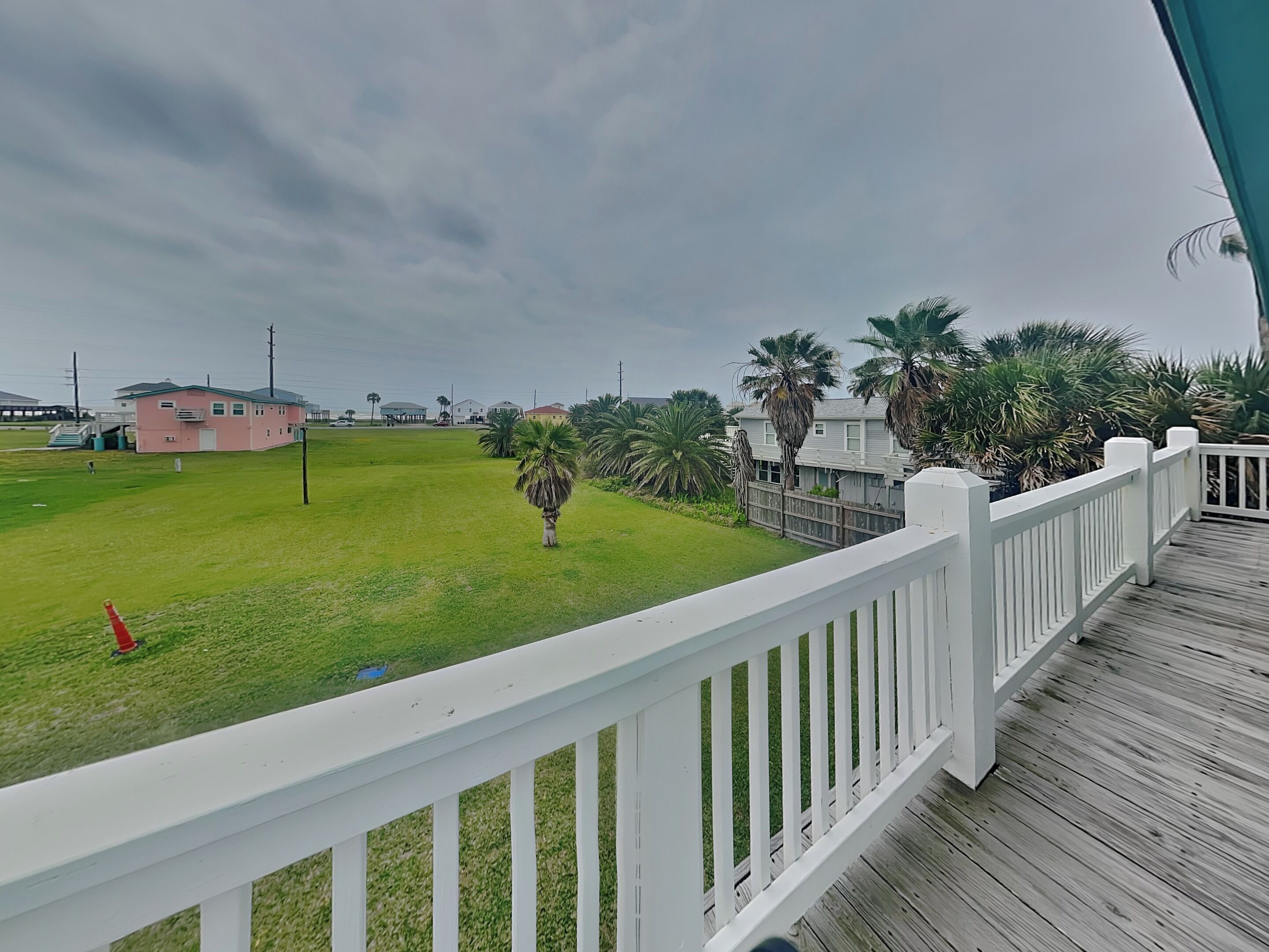 A wooden deck with a white railing, a grassy area with a red cone, and palm trees.