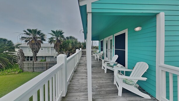 A wooden deck with white railings, a turquoise building, and several white Adirondack chairs. - Sundancers (Galveston)