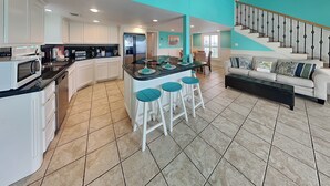 A modern kitchen with white cabinetry, a central island with stools, and a staircase leading to a living area with a sofa and cushions. - Sundancers (Galveston)