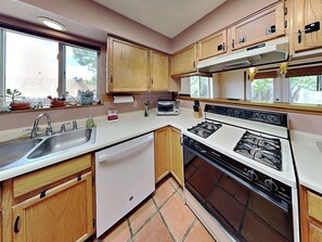 A compact kitchen with wooden cabinets, a white countertop, a gas stove, a dishwasher, and a window providing natural light.