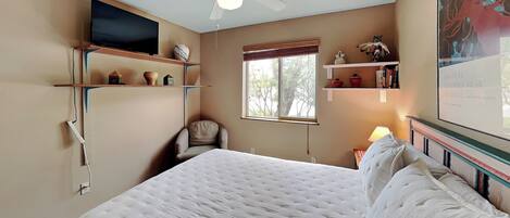 A bedroom with a bed, a ceiling fan, a TV mounted on the wall, shelves with decorative items, a window with a view, and a framed poster on the wall.