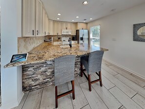 A kitchen with a granite countertop, two bar stools, a microwave, a refrigerator, and a stone backsplash. - Jamaica Beach-birds of Paradise (Galveston)