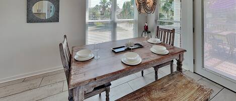 Una mesa de comedor con platos y vasos blancos, un banco, una lámpara colgante y vistas a palmeras a través de las ventanas.