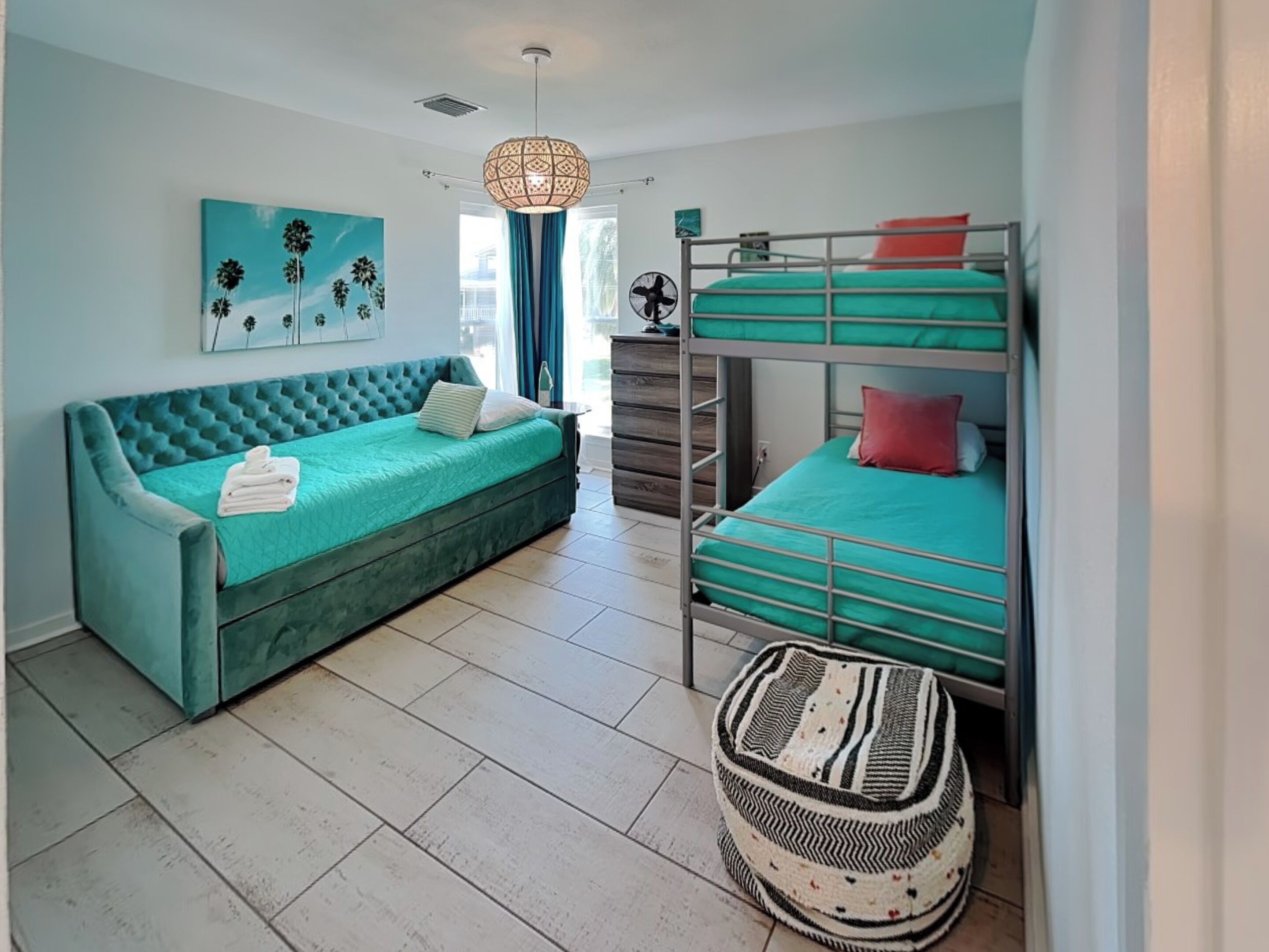 A room with a teal sofa, bunk beds, a woven ottoman, a hanging light fixture, and a painting on the wall.