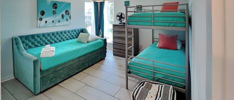 A room with a teal sofa, bunk beds, a woven ottoman, a hanging light fixture, and a painting on the wall.