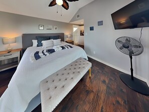 A bedroom with a bed, bedside tables, a bench, a ceiling fan, and a TV mounted on the wall. - Jamaica Beach-birds of Paradise (Galveston)