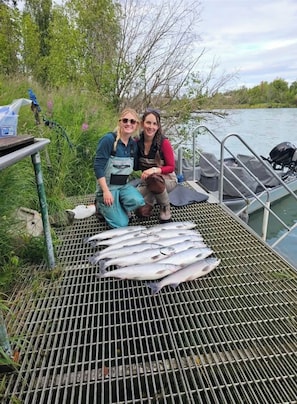 Zwei Personen auf einem Boot, das an einem Fluss vertäut, mit einem Grilltisch und Fisch darauf.