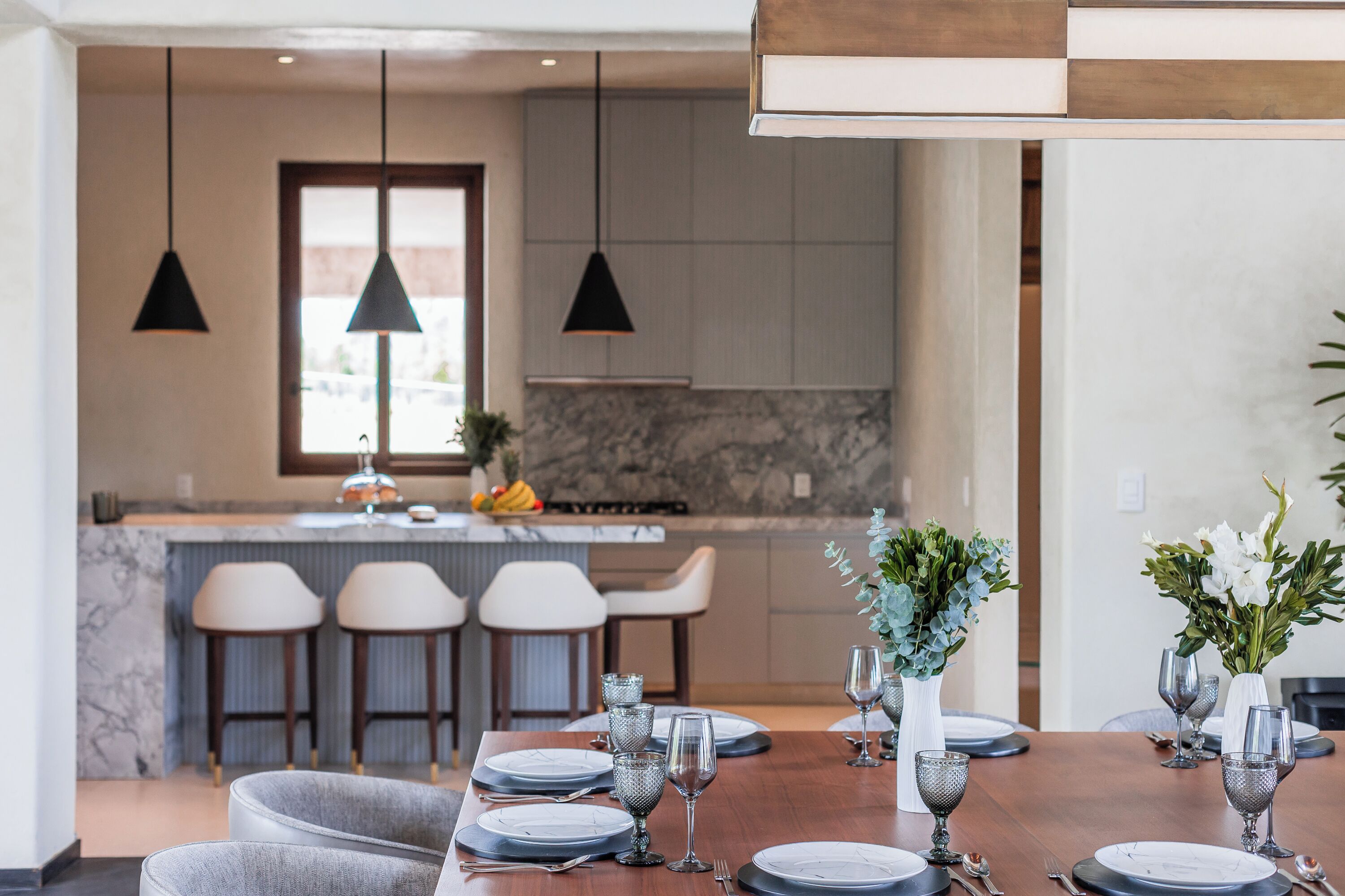 A modern dining area with a long table set for a meal, a kitchen in the background, and pendant lighting.