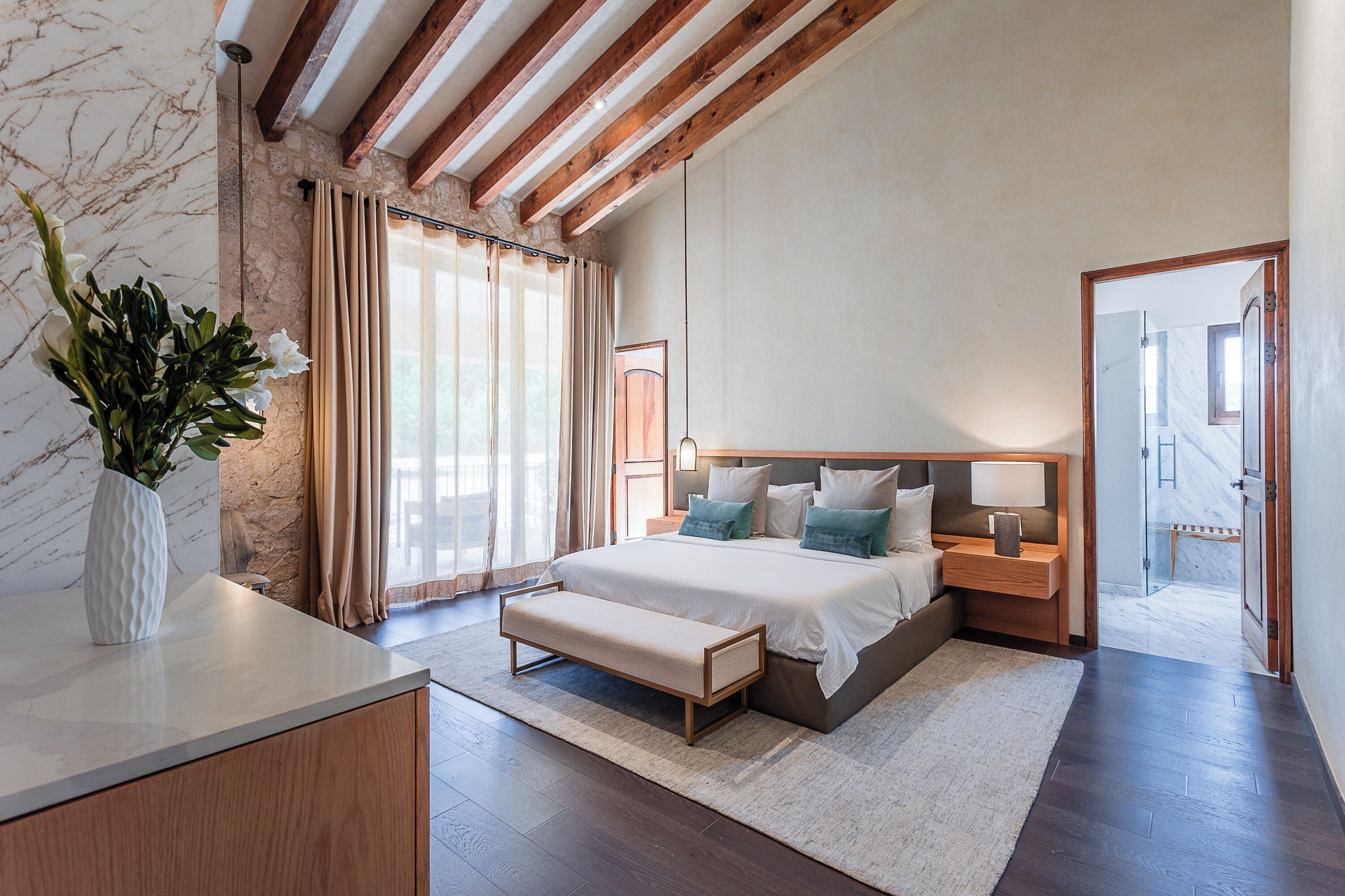 A bedroom with a bed, bedside tables, a bench, a vase with flowers, and a view of a hallway.