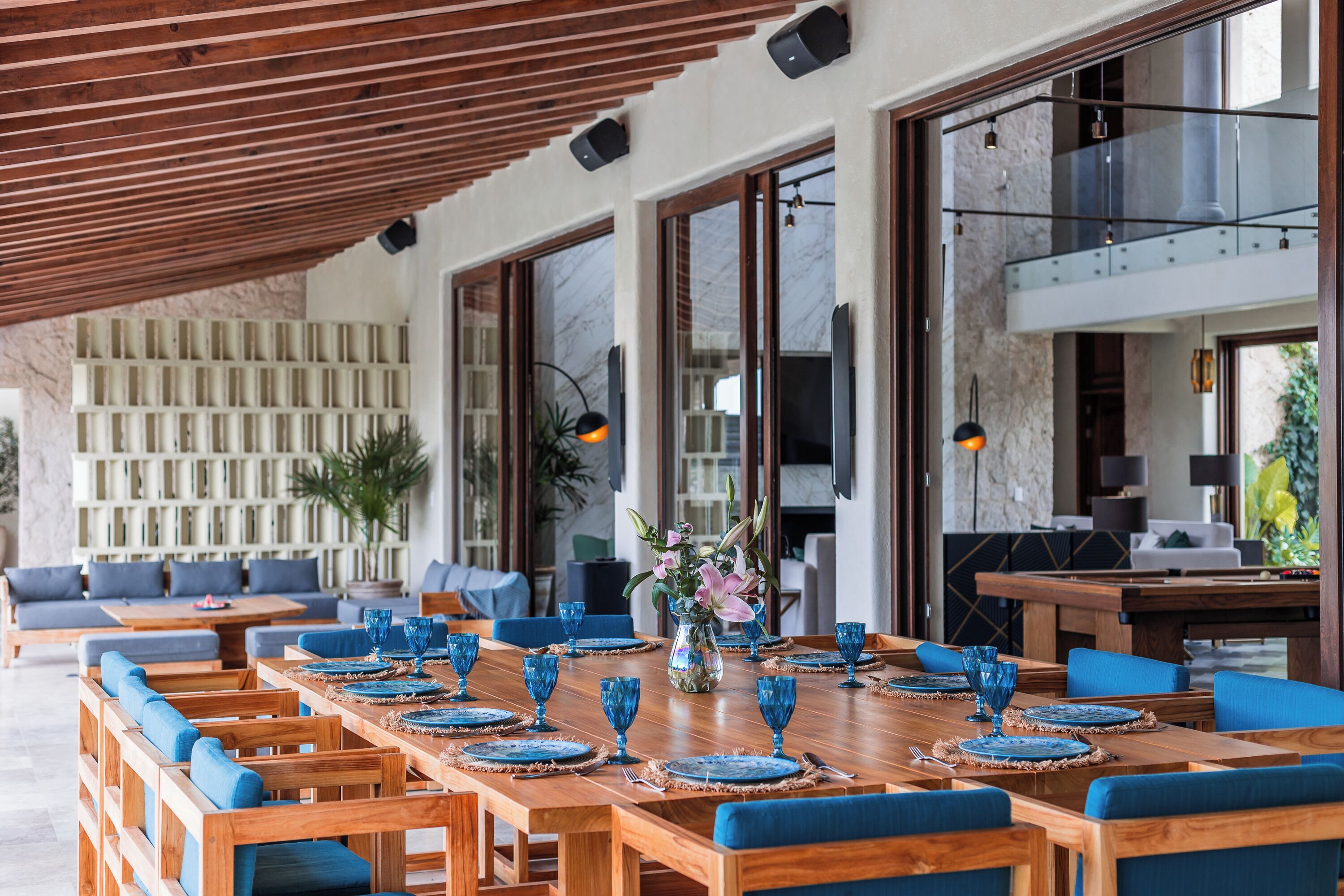 A spacious outdoor dining area with wooden tables and blue upholstered chairs, set against a backdrop of a stone wall and large windows.