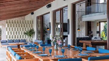 A spacious outdoor dining area with wooden tables and blue upholstered chairs, set against a backdrop of a stone wall and large windows.