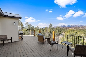Eine Holzterrasse mit Gartenmöbeln, einem Grill und Blick auf Berge und Bäume.