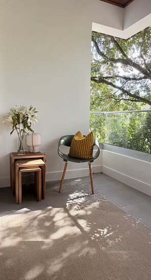 Ein Zimmer mit Blick auf Bäume, ein Sessel mit gelber Sitzkissen, ein kleiner Holztisch mit Blumenvase und ein gewebter Teppich.