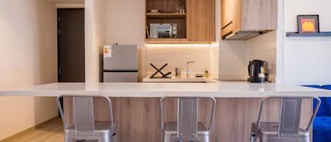 A modern kitchen with a white countertop, stainless steel appliances, and wooden cabinets. There are three metal bar stools at the counter.
