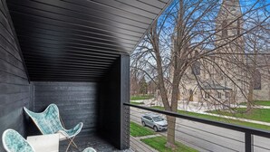 A balcony with two chairs and a table, overlooking a street and a building. - Two Private Spaces | Downtown & Near Beach (Sheboygan)