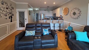 A living room with a black leather sofa, a ceiling fan, and a kitchen in the background. - Coastal Retreat w/ Balcony, Bikes & Pool Access (New Bern)