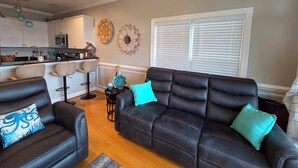 A living room with a black leather sofa, two bar stools, a kitchen in the background, and a decorative wall piece. - Coastal Retreat w/ Balcony, Bikes & Pool Access (New Bern)