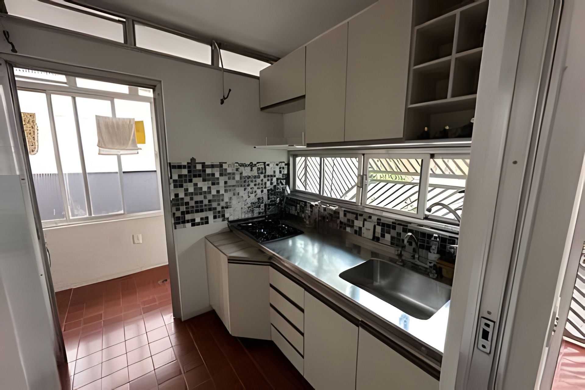 Cozinha compacta com armários brancos, bancada preta e uma janela com grade de metal.