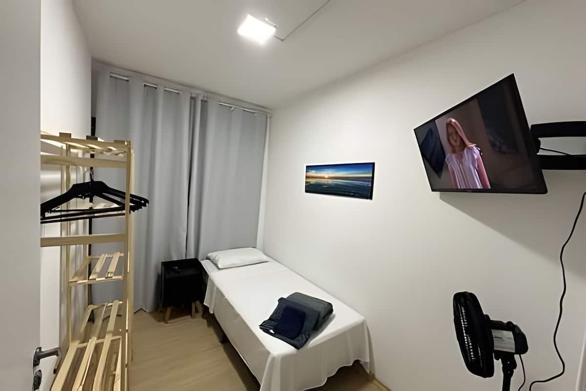 Ein kleines Zimmer mit einem Einzelbett, einem Fernseher, einem Wandbild, einem Ventilator und einem hölzernen Kleiderständer.