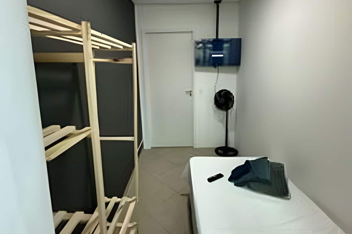 Ein schmales Gangzimmer mit Etagenbett, einer weißen Tür, einem an der Wand montierten Fernseher und einem Standventilator.