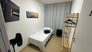 Quarto pequeno e arrumado com uma cama de solteiro, TV na parede, janela com cortinas e um porta-roupas de madeira com cabides.