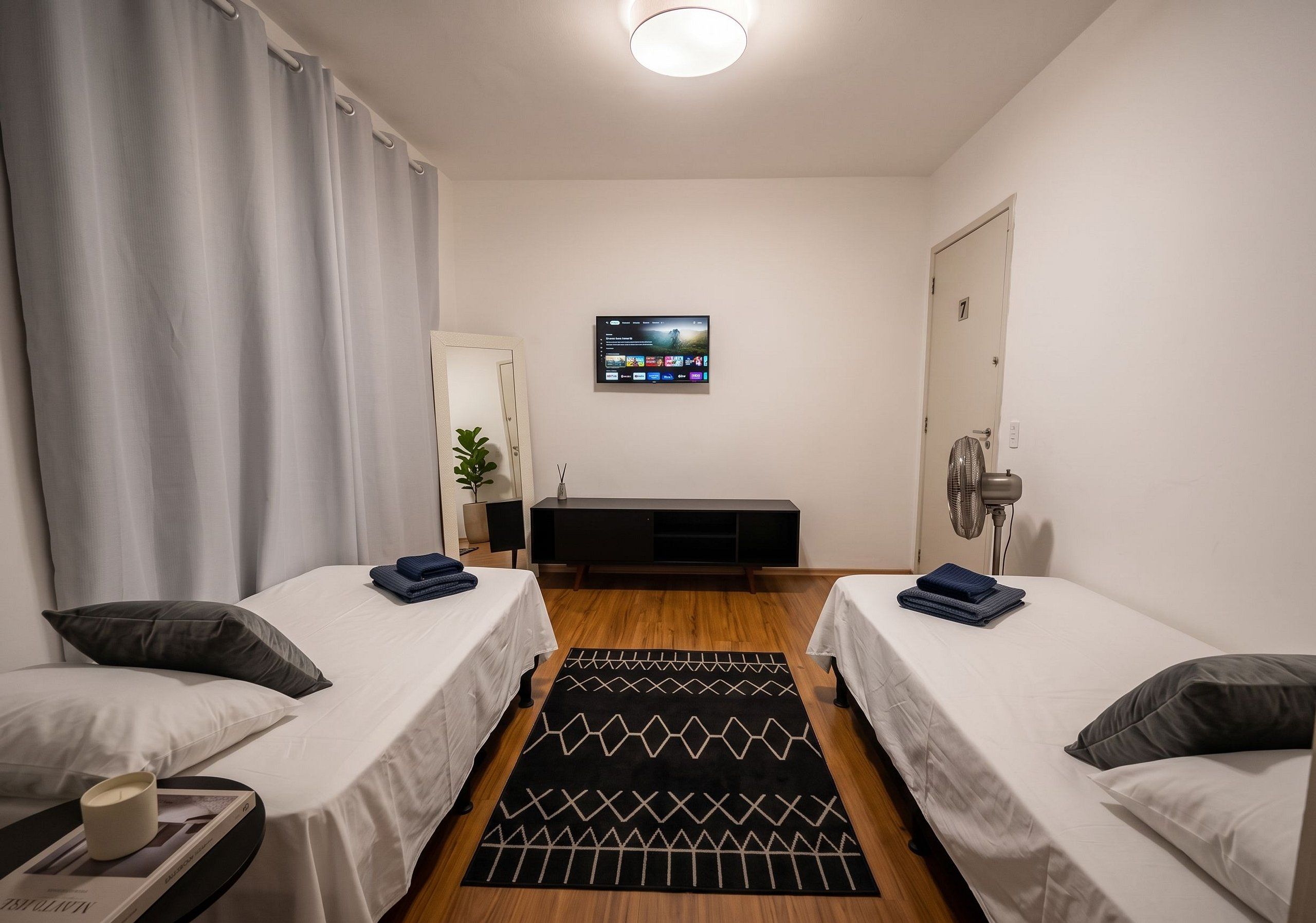 A hotel room with two single beds, a TV mounted on the wall, a bedside table with a lamp, a small potted plant, and a black and white patterned rug.