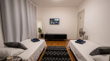 Habitación de hotel con dos camas individuales, un televisor montado en la pared, una mesita de noche con lámpara, una pequeña planta en maceta y una alfombra con estampado blanco y negro.