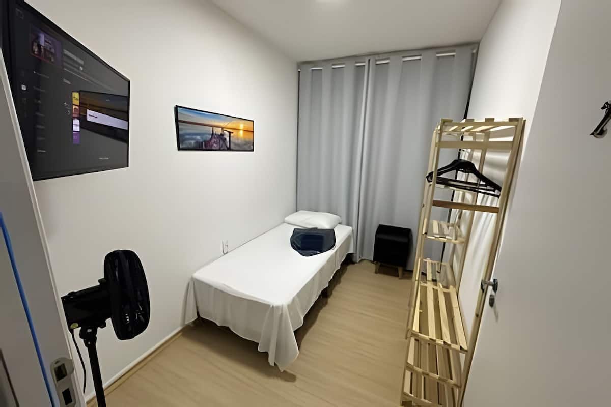 Ein kleiner, ordentlicher Raum mit einem Einzelbett, einem an der Wand montierten Fernseher, einem Fenster mit Vorhängen und einem Holzgestell mit Kleiderhaken.