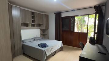 Quarto com cama, escrivaninha, cadeira e janela com vista para o verde.