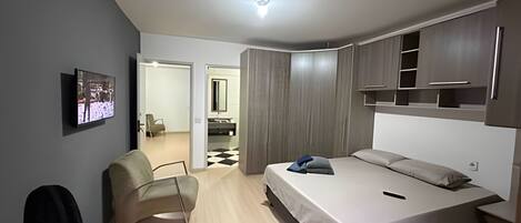 Un dormitorio moderno con cama, una silla, televisor y armario.