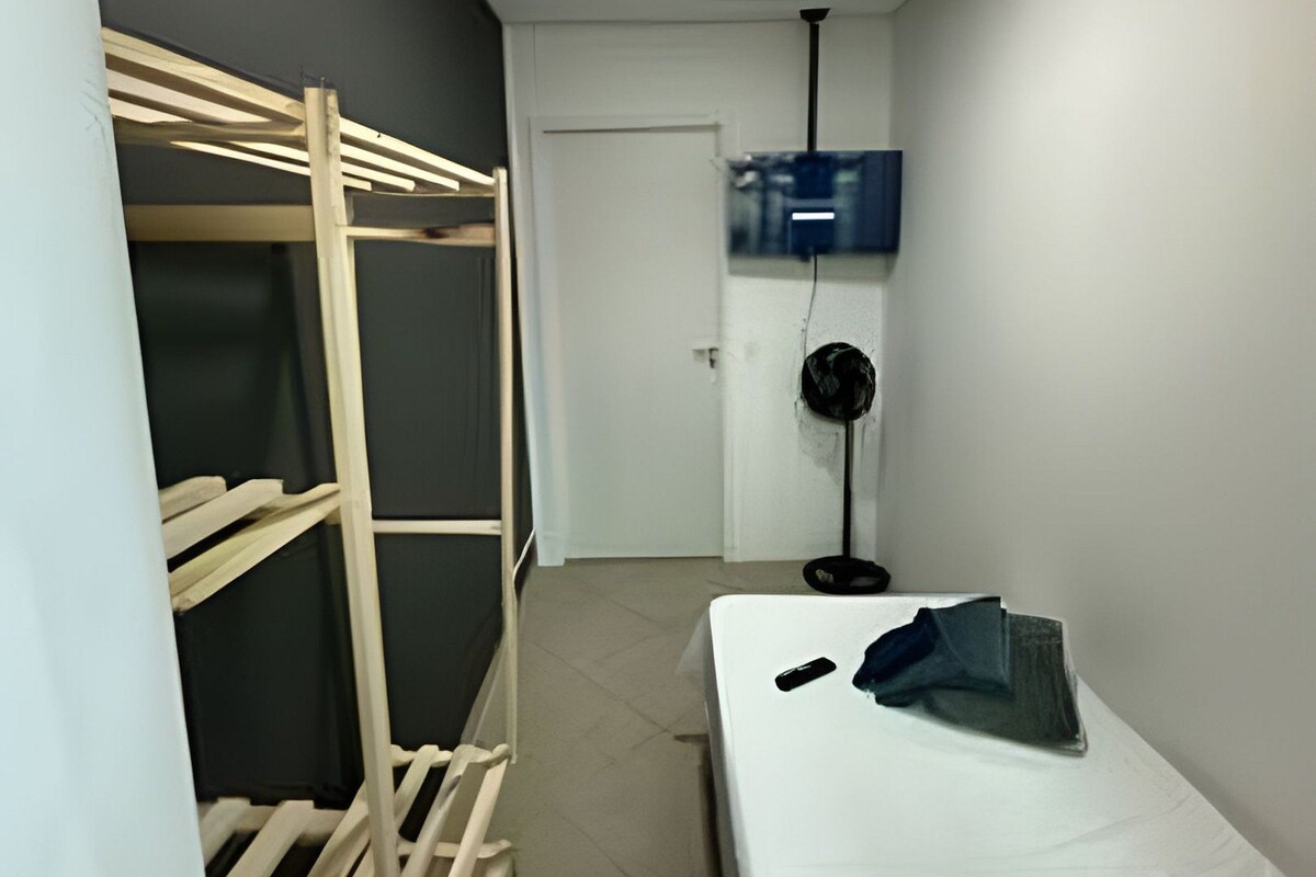 A narrow hallway with a bunk bed, a white door, a wall-mounted TV, and a standing fan.