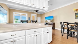 A kitchen with white cabinetry, a dining area with a table and chairs, a ceiling fan, and a painting on the wall. - Casa Valencia (Panama City Beach)