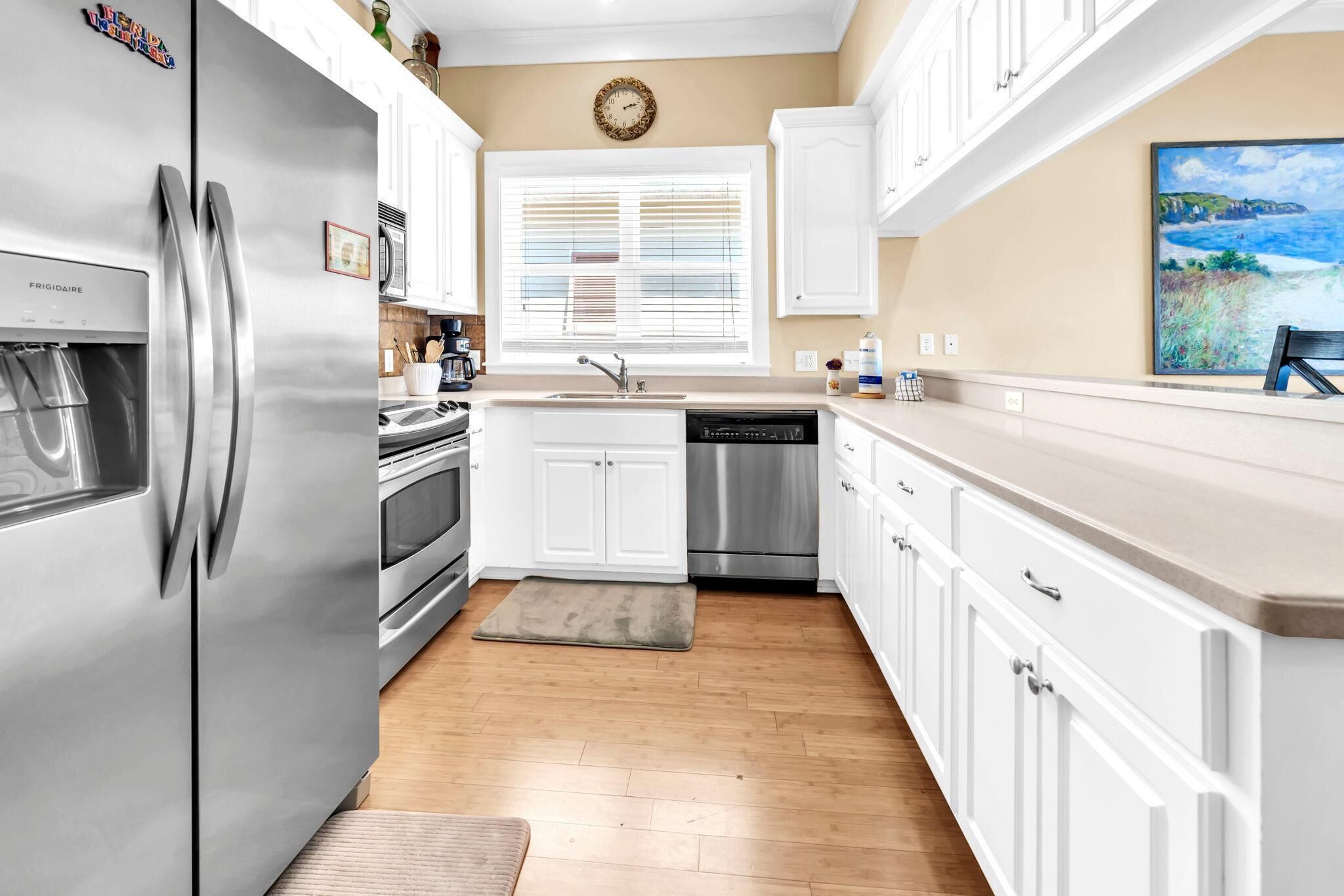 A kitchen with white cabinets, a stainless steel refrigerator, a dishwasher, and a window with blinds.