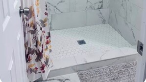 A shower with a hexagonal tile pattern, a small shelf with toiletries, and a door with a floral curtain.