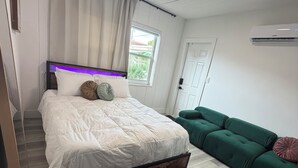 A bedroom with a bed, a green sofa, a window, and an air conditioning unit.