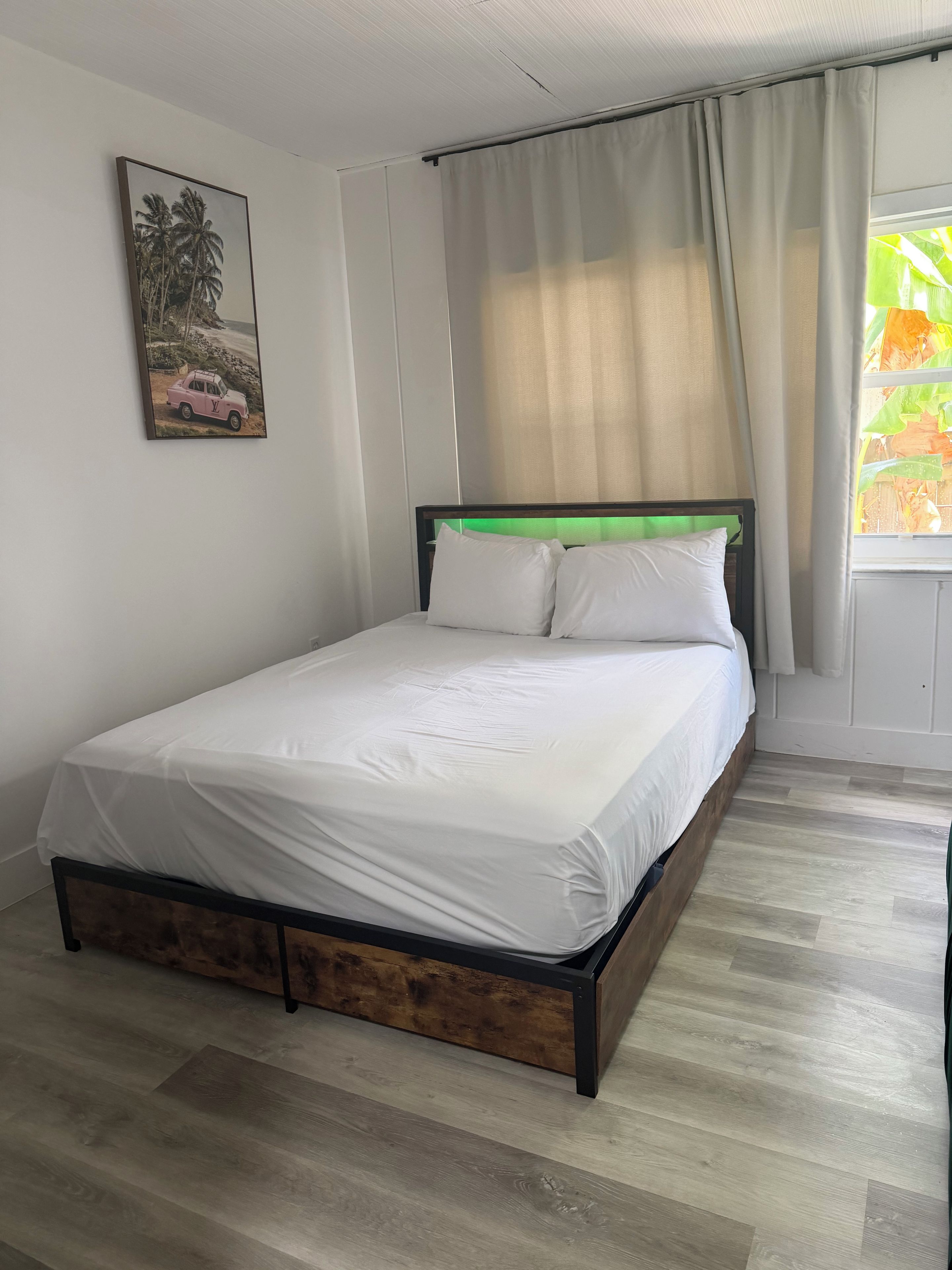 Un lit bien fait, avec du linge de lit blanc, dans une pièce au plancher en bois, ornée d’un tableau au mur et dotée d’une fenêtre avec des rideaux.