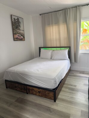 A neatly made bed with white linens in a room with wooden flooring, a painting on the wall, and a window with curtains.