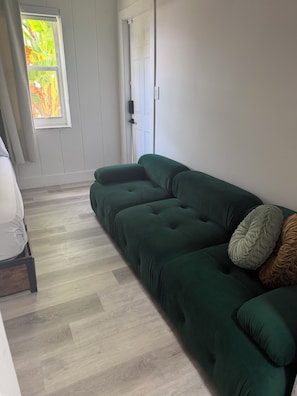 A room with a green sofa, a white chair, and a window with a view of greenery.