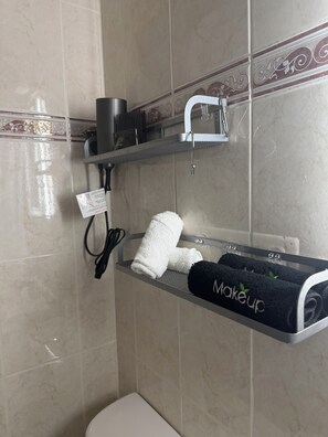A bathroom with a towel rack holding a white towel and a black towel with 'makeup' written on it. There's a shelf above the rack with a hairdryer and other toiletries.