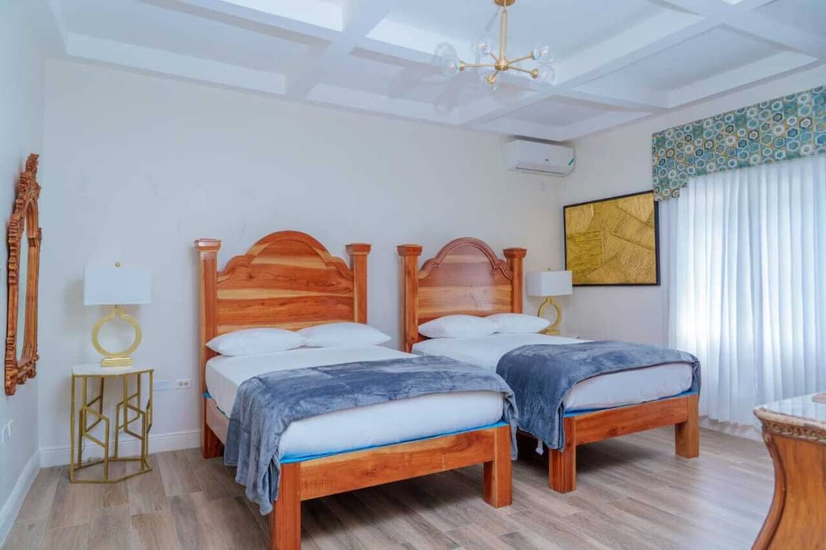 A double room with two wooden beds, a chandelier, a large window with curtains, and a wall-mounted air conditioner.
