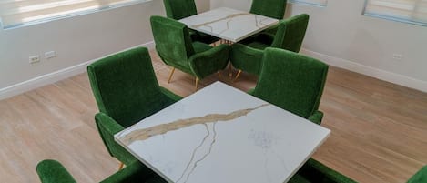 Een kamer met een witte tafel en groene stoelen, grote ramen met luxaflex en een vloer in een lichte kleur.