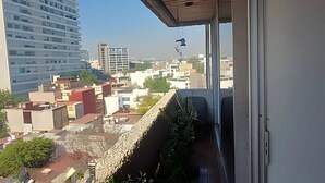Un balcón con vistas a edificios circundantes, una planta colgante y una ventana con cortina.