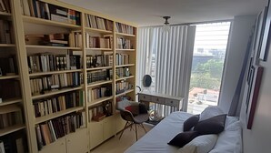 Un dormitorio con una amplia estantería, un escritorio con silla, una cama con almohadas y una ventana con cortinas.