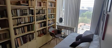 Ein Schlafzimmer mit einem großen Bücherregal, einem Schreibtisch mit Stuhl, einem Bett mit Kissen und einem Fenster mit Vorhängen.