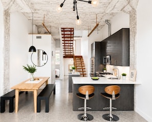 Una cocina moderna con una isla central, taburetes de bar, un banco de madera y una escalera que sube a un nivel superior.