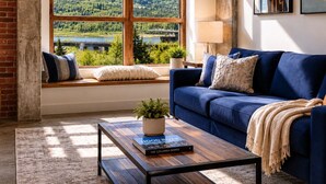 Une pièce avec une grande fenêtre, un canapé bleu, une table basse avec un livre et une lampe.
