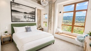 Une chambre avec un grand lit, une vue sur l’extérieur, un coin fenêtre aménagé en siège et un tableau accroché au mur.