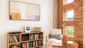 Une pièce avec un mur en briques, une bibliothèque remplie de livres et un tourne-disque, un fauteuil avec un coussin, et un grand tableau abstrait accroché au mur.