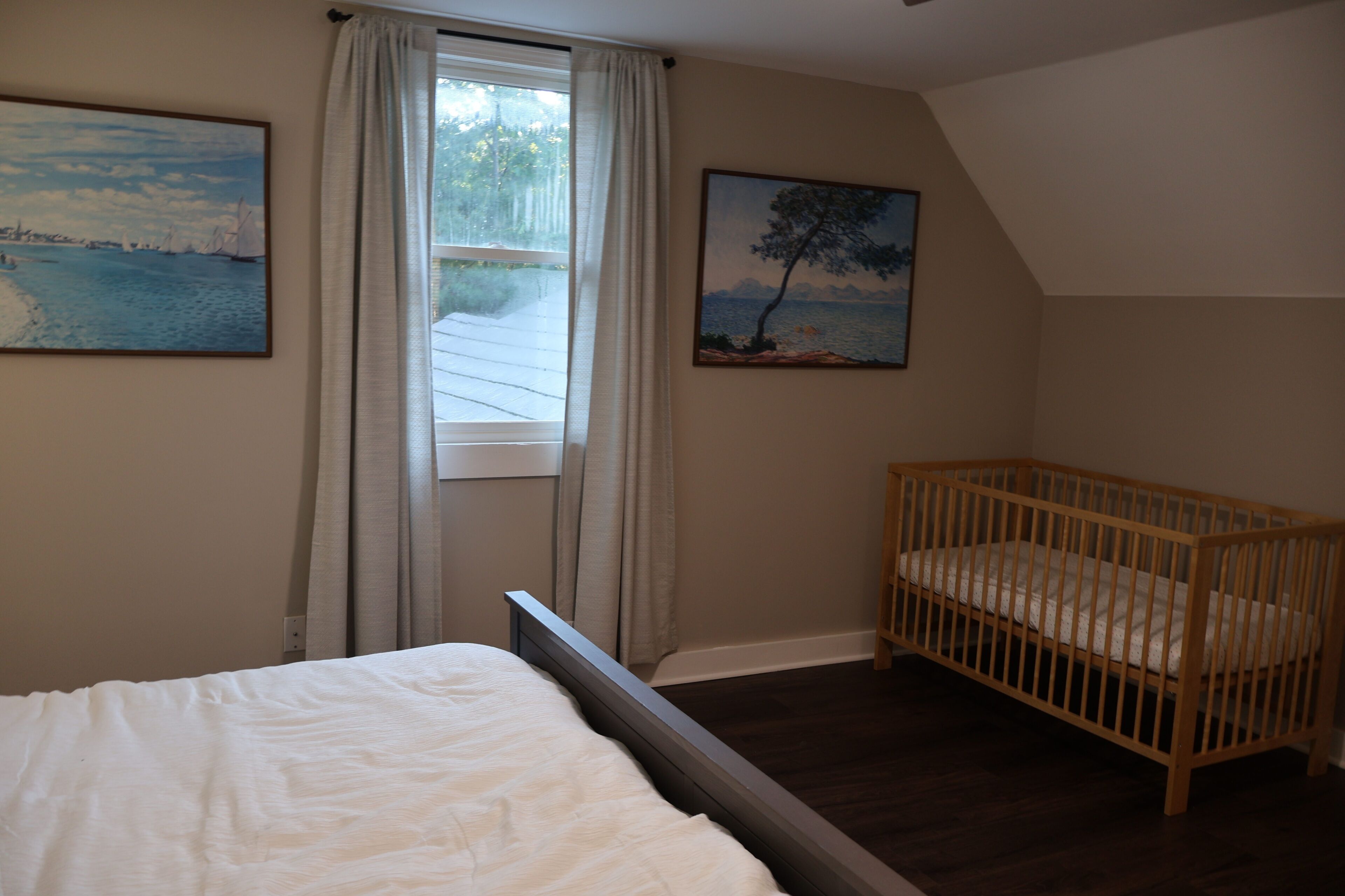 A bedroom with a bed, a crib, a window with curtains, and two paintings on the wall.