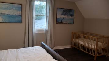 A bedroom with a bed, a crib, a window with curtains, and two paintings on the wall.